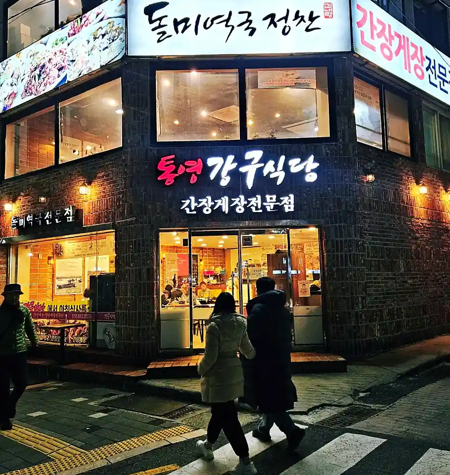 한국인 30대 남녀가 통영 식당에서 굴구이를 집게로 집어 올리는 실사용 장면, 배경에는 한국어 메뉴판이 보임